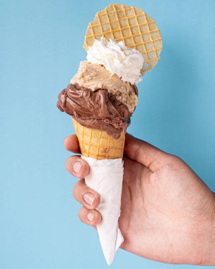 Çikolatalı dondurma, fındık ezmeli ve kremalı nefis waffle külahı, yanında da bir eliyle mavi arka planda tutulan waffle kurabiyesi..