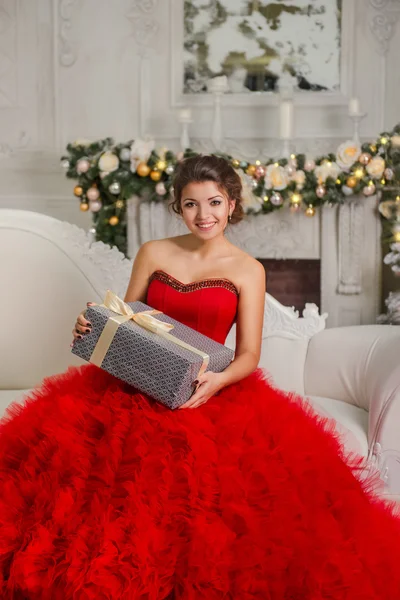 Piękna, uśmiechnięta brunetka siedzi na kanapie, trzymając prezent na Boże Narodzenie — Zdjęcie stockowe