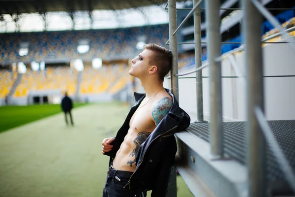 Joven con chaqueta en el pecho desnudo, de pie en el estadio, con las barandillas — Foto de Stock