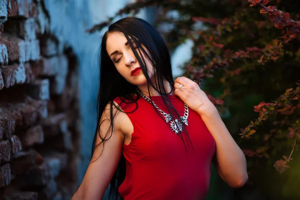 Retrato de beleza de moda. Mulher bonita com cabelo encaracolado vestindo um vestido vermelho moda, de pé contra a parede, fechou os olhos — Fotografia de Stock