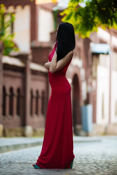 Krásná mladá brunetka v červených šatech pózuje v ulici nedaleko červené cihlové zdi — Stock fotografie
