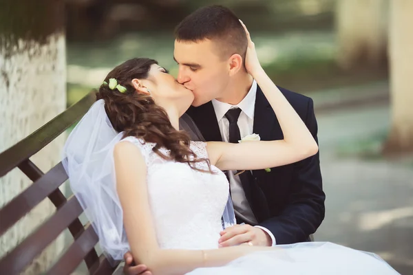 Aantrekkelijke brunette bruid in elegante jurk en knappe bruidegom in blauwe pak op huwelijksdag heerlijk knuffelen op de Bank buiten. Kersverse vrouw en man omarmen in groen park. Lief bruidspaar — Stockfoto