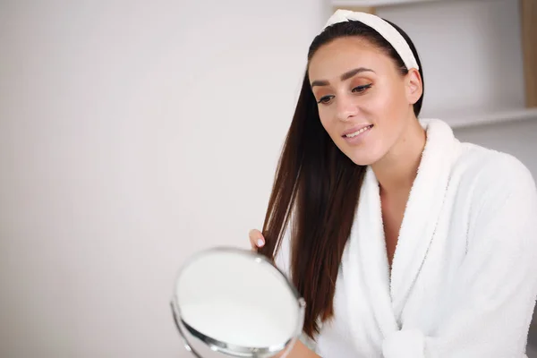 Красота, уход за кожей и концепция людей - улыбающаяся молодая женщина в повязке для волос, касающаяся своего лица и смотрящая в зеркало в домашней ванной комнате — стоковое фото