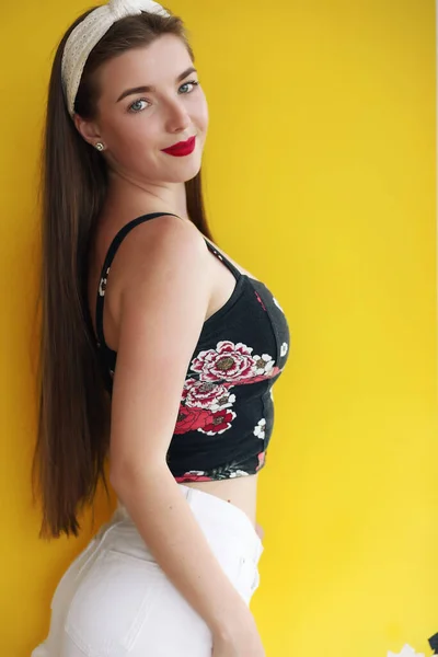 Happy girl in vintage denim shorts standing in confident pose on yellow background. Indoor portrait of enchanting young lady in summer casual outfit plays with her hair. — Stock Photo, Image