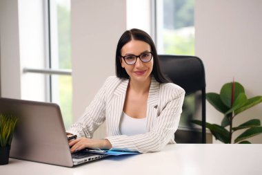 Önemli bir belgeye sahip neşeli esmer kadın. Dostane gülümseyen ve bilgisayarını kontrol ederken kameraya bakan.