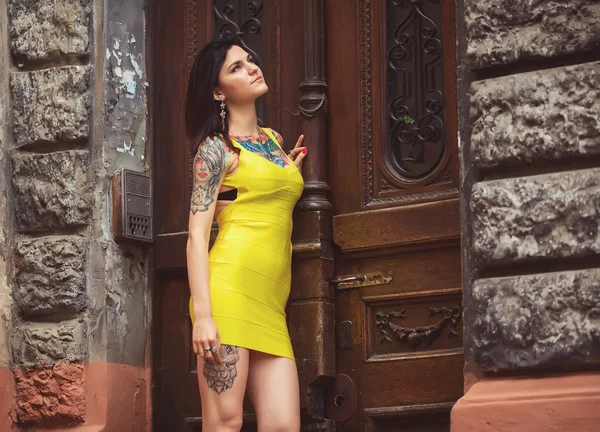 Красивая женщина в жёлтом платье, татуировки, опирающаяся на старую дверь — стоковое фото