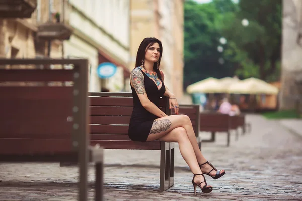 Beautiful girl in a black dress sitting on a bench, covered in t — Stock Photo, Image