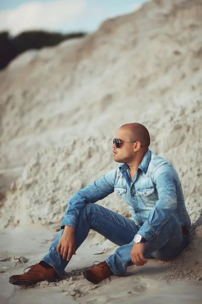 Chico sentado en la arena, una ropa de jeans bronceados — Foto de Stock