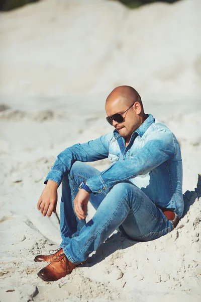 Hombre con estilo en jeans sentados en la arena, con gafas — Foto de Stock