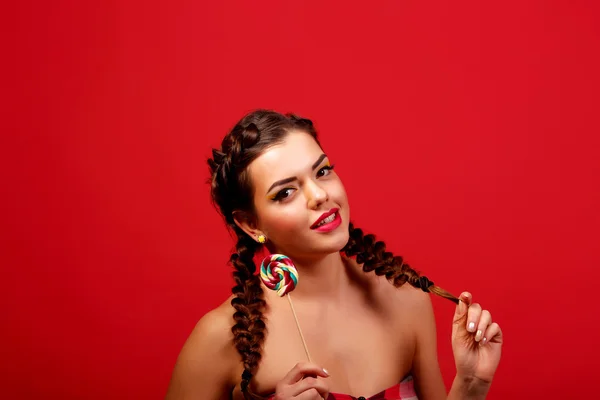 Güzellik moda modeli kız renkli lolipop yemek. Sürpriz komik genç kadınla saç modeli, çivi ve makyaj bej, kırmızı bir arka plan üzerinde izole örgü