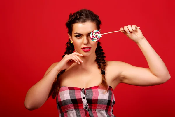 Belleza modelo chica comer piruleta colorido. Mujer joven divertida sorprendida con trenzas peinado, uñas y maquillaje beige, aislado sobre un fondo rojo —  Fotos de Stock