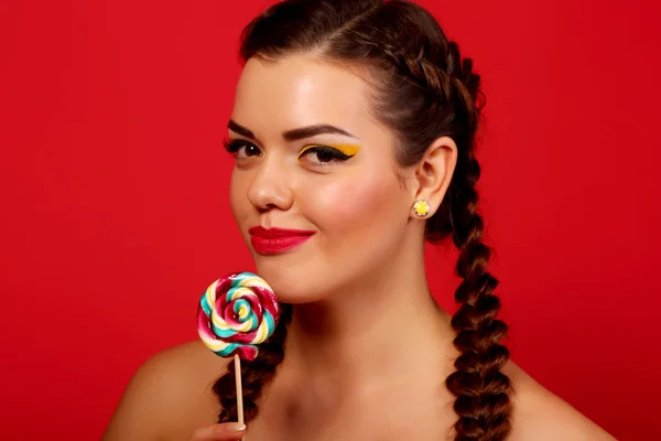 Uroda moda model dziewczynka jedzenie kolorowy lizak. Zaskoczony śmieszne młoda kobieta z plecionki fryzury, paznokci i makijażu beżowy, na białym tle na czerwonym tle — Zdjęcie stockowe