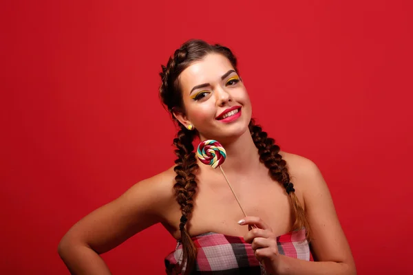 Skönhet mode modell flicka äter färgglada lollipop. Förvånad rolig ung kvinna med flätor frisyr, naglar och smink beige, isolerad på en röd bakgrund — Stockfoto