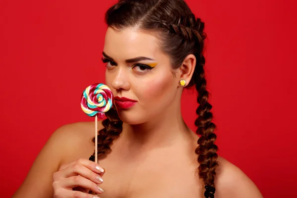 Beauty fashion model girl eating colorful lollipop. Surprised funny young woman with braids hairstyle, nails and makeup beige, isolated on a red background — Stock Photo, Image