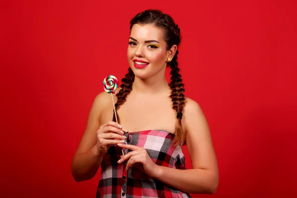 Funny young girl holding a lollipop and posing for kamer? over red background, red background, pigtails — Stock Photo, Image