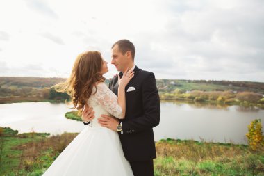 Beautiful young couple wedding guy in blue jacket covering brunette in a white dress with red lips holding bouquet of flowers, standing on the sun background, horizontal image