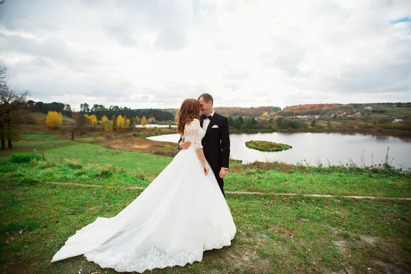 Matrimonio, bacio di coppia sulle colline vicino al fiume al tramonto. Vento svolazzante un lungo velo. Paesaggio di colline e montagne — Foto Stock