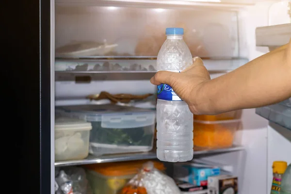 Woman opening refrigerator door with bottle of fresh water near open fridge, closeu