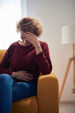 Karın ağrısı ve baş ağrısı olan bir kadın evdeki kanepede oturuyor..