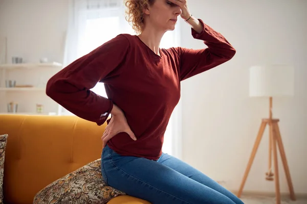 Woman with head and hip pain sitting on a couch at home.