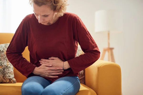Woman with stomach pain problem sitting on a couch at home.