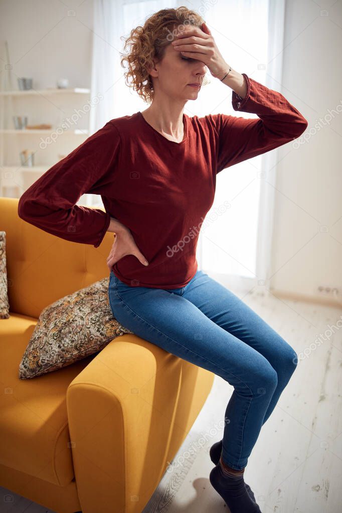 Woman with head and hip pain sitting on a couch at home.