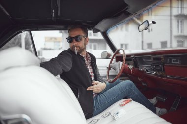 Elinde sigara tutan ve klasik Amerikan arabası süren retro giysili bir adam..
