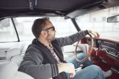 Elinde sigara tutan ve klasik Amerikan arabası süren retro giysili bir adam..