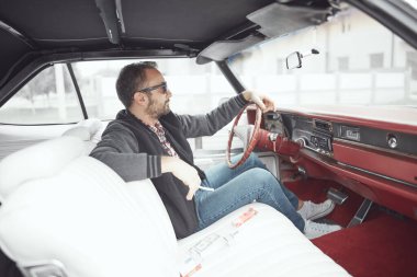 Elinde sigara tutan ve klasik Amerikan arabası süren retro giysili bir adam..