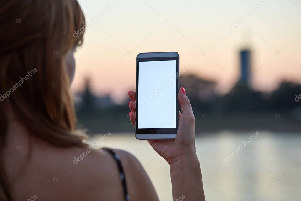 Young woman holding cellphone with blank white screen in urban city surroundings.