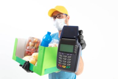 Beyaz bir duvarın önündeki teslimatçı. Koruyucu maskeli. Yanında bakkaliye ve POS var..