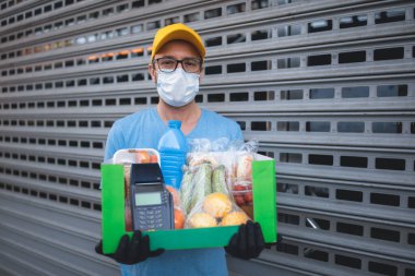 Binanın önündeki koruyucu maskeli teslimatçı bakkaliye ve POS ile temas kurulamayan ödeme kutusuyla..