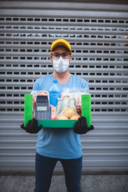 Binanın önündeki koruyucu maskeli teslimatçı bakkaliye ve POS ile temas kurulamayan ödeme kutusuyla..