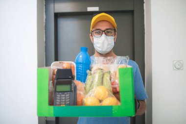 İnşaat asansörünün önündeki teslimatçı. Koruyucu maske takıyor. Yanında bakkaliye ve ulaşılmayan ödemeler için POS var..