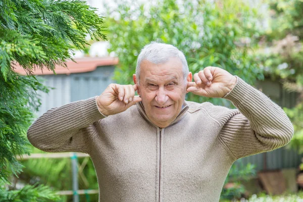 Elderly Caucasian in green garden covers his ears with his fingers not to hear