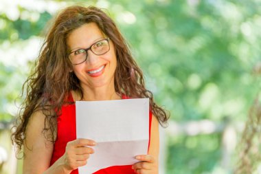 gorgeous middle-aged woman in a red dress and long brown wavy hair is reading a paper and smiling while wearing a pair of nerdy eyeglasses in a garden clipart
