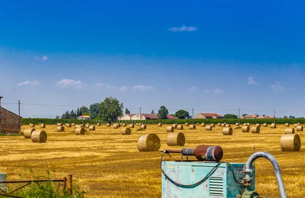 round hay bales in a harvested field near a country farmhouse and pump for irrigation in the foreground, rural and bucolic atmosphere of a hot summer day