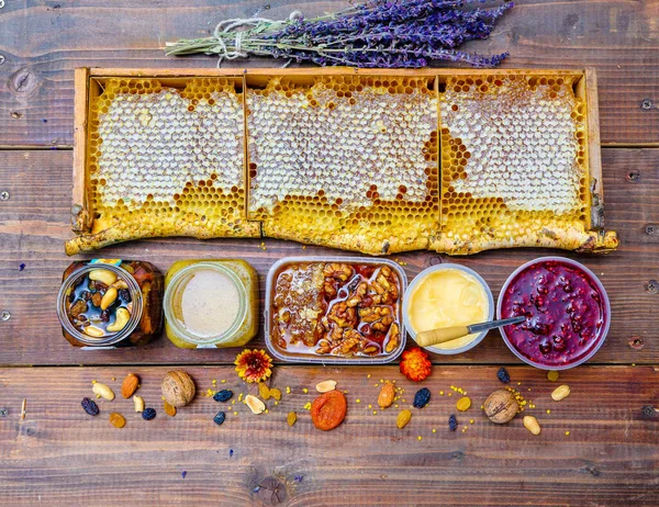 養蜂製品との素朴な静物画 ガラス瓶の中の蜂蜜の櫛 白樺の枝のハニカムフレームの野生の蜂蜜 ラズベリージャム 蜂蜜のスフレ ドライフルーツやナッツ 木製の背景に — ストック写真