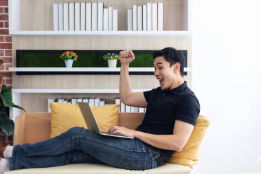 Kanepede oturan bilgisayarlı dizüstü bilgisayar kullanan adam mutlu bir ifade vermek için ellerini kaldır.