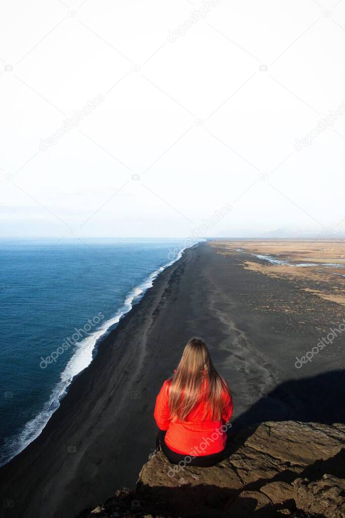 Red jacket female tourist sitting on rock cliff atlantic ocean coast Dyrholaey black sand volcanic beach Vik Iceland