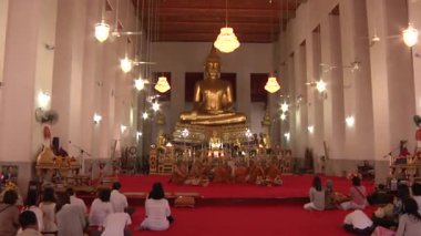 Bangkok 'ta. Tayland. Tapınaktaki insanlar keşişlerle birlikte Buda heykelinin önünde oturuyorlar. Kamera yakınlaşıyor ve Buda 'nın yüzünü yakına çekiyor. 