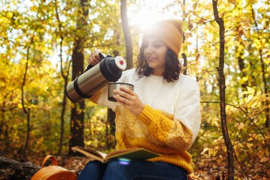 Turuncu şapkalı, kazaklı ve sırt çantalı genç bir kadın sonbahar ormanındaki termostan çay döküyor. Sonbaharda kadın turist parkta yürüyüş yapıyor ve ağaçların arasında oturmuş gülüyor.