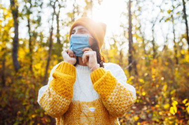 Turuncu şapka ve kazak giyen genç bir kadın sonbahar ormanında tıbbi steril maskeyi çıkarıyor. Kadın sonbahar parkında Coronavirus covid-19 salgını sırasında temiz hava soluyor. Sağlık hizmetleri kavramı