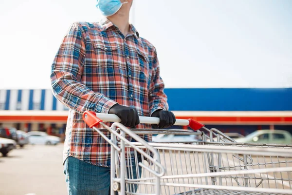 Young man stands outside the supermarket with the trolley wearing sterile medical mask and gloves to protect from coronavirus. Customer at a shops parking is going for purchases. Healthcare concept