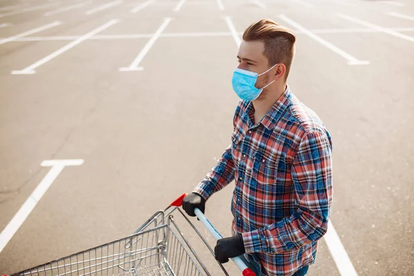 Young man stands outside the supermarket with the trolley wearing sterile medical mask and gloves to protect from coronavirus. Customer at a shops parking is going for purchases. Healthcare concept