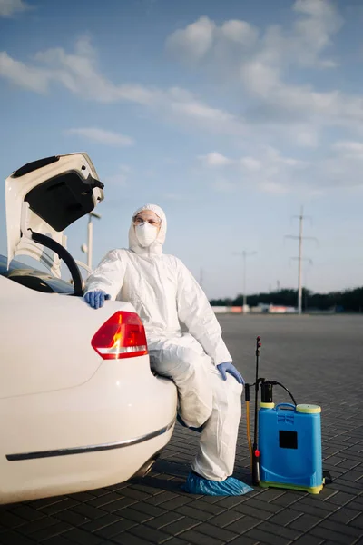 Disinfection service worker sits on a trunk of a white car with a spray equipment near his leg. Sanitizing professional wearing protective suit, mask and gloves in the vehicle. Coronavirus prevention