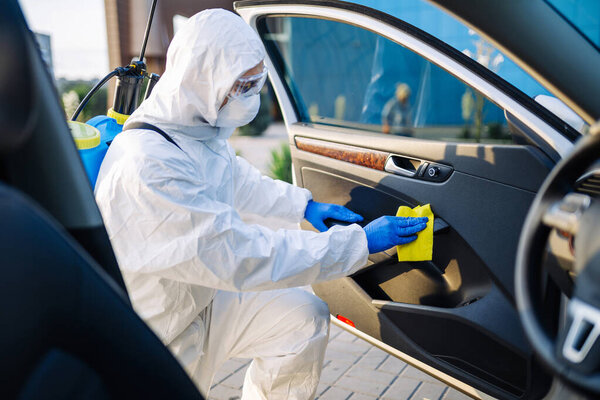 Sanitizing service worker cleans the car's interior with a yellow rug. A man in a protective suit, mask and gloves disinfects the vehicle's doors. Coronavirus covid spread prevention concept