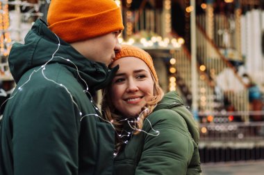 Loving young couple standing together outdoors among garlands at winter season. A boy and a girl having fun, hug and kiss each other in the lights of Christmas market. Holidays concept