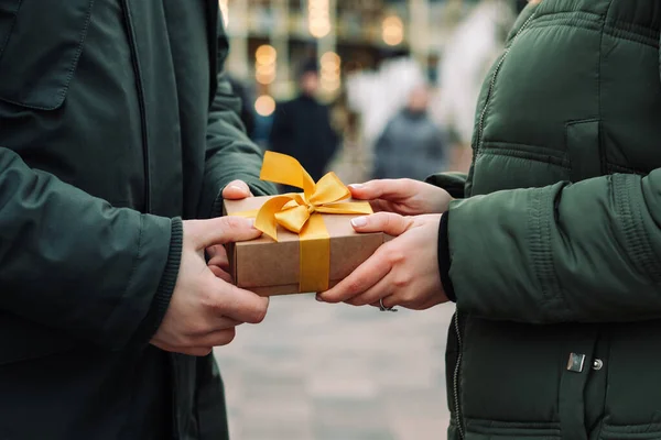 Hands of a couple with a gift present. Man gives a small present with a yellow ribbon to his girlfriend on the street. Woman takes a gift from her boyfriend. Valentines day and Christmas concept