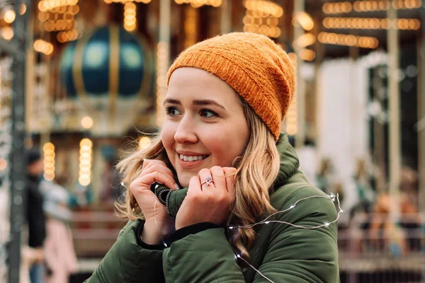 Young woman's portrait at a Christmas market decorated with lights. Female wearing orange hat and green coat stands at a seasonal New Year market. Winter mood concept